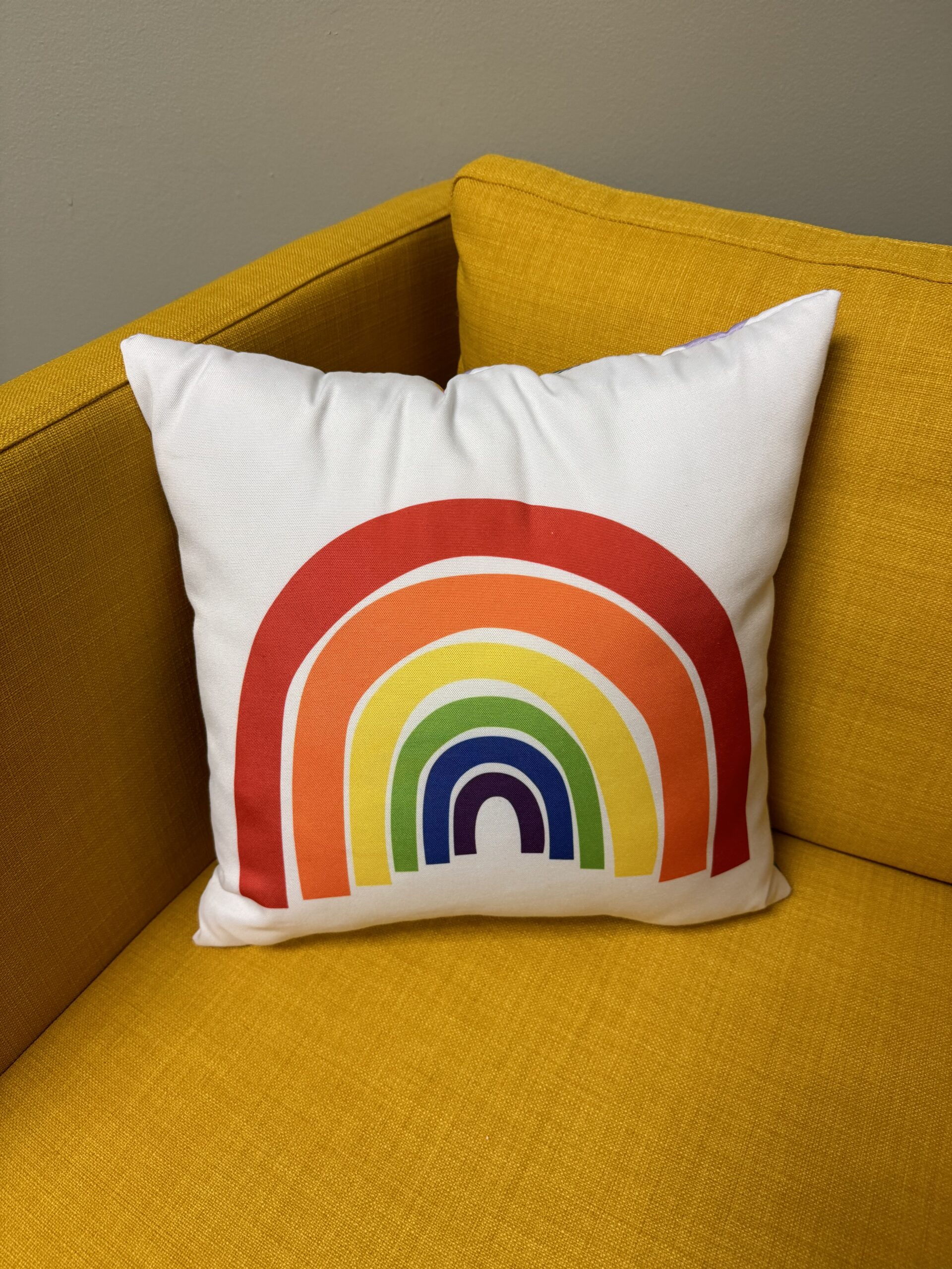 A white throw pillow with a rainbow design placed on a yellow couch.