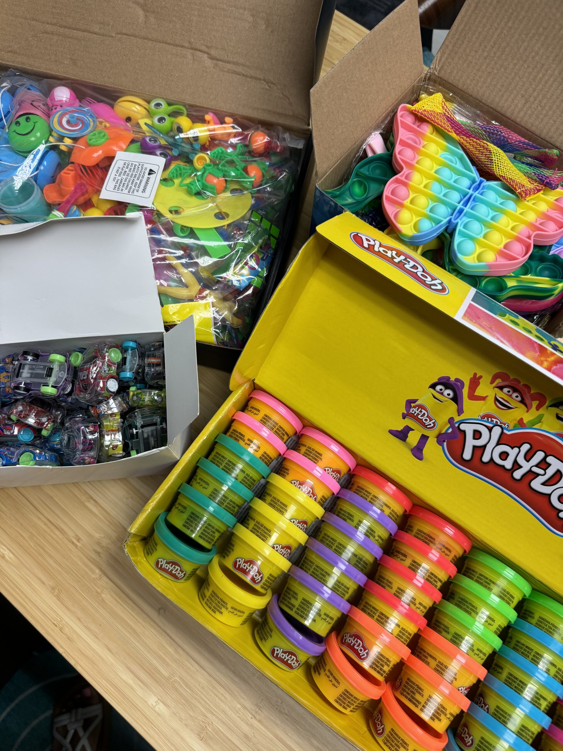 A collection of colorful sensory toys, including Play-Doh containers, fidget spinners, and pop-it toys, displayed in open boxes on a wooden table.