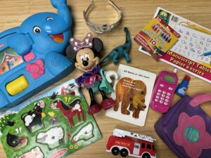 A collection of children's toys and educational materials spread out on a wooden surface, including a toy elephant, Minnie Mouse doll, book, puzzle, toy fire truck, play money, tiara, and more.